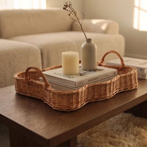 May include: A light brown woven wicker tray with a scalloped edge and two arched handles. Inside the tray are two stacked books with the word DESIGN printed on the spine, a cream-colored pillar candle, and a small ceramic vase with a dried twig.