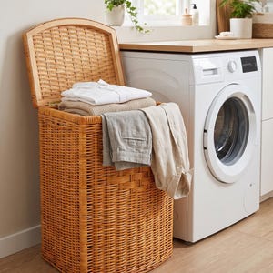 Puede incluir: Un cesto para la ropa alto y rectangular de mimbre marrón claro con tapa abatible. El cesto está lleno de ropa doblada blanca y beige, que incluye una camisa blanca, un suéter beige, pantalones grises y una camisa beige colgada por el borde.