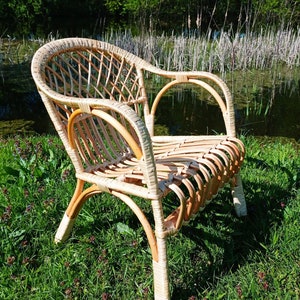May include: A wicker chair with a light brown finish sits on a bed of green grass. The chair has a woven back and seat, and the arms are curved. The chair is in a natural setting, with green grass and trees in the background.