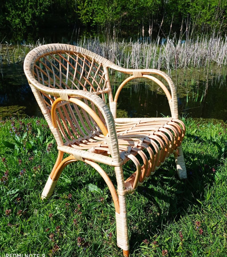 May include: A wicker chair with a light brown finish sits on a bed of green grass. The chair has a woven back and seat, and the arms are curved. The chair is in a natural setting, with green grass and trees in the background.
