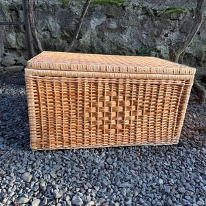 May include: A rectangular wicker storage basket with a lid. The basket is made of woven light brown wicker and sits on a bed of gray gravel. The basket has a woven pattern on the front.