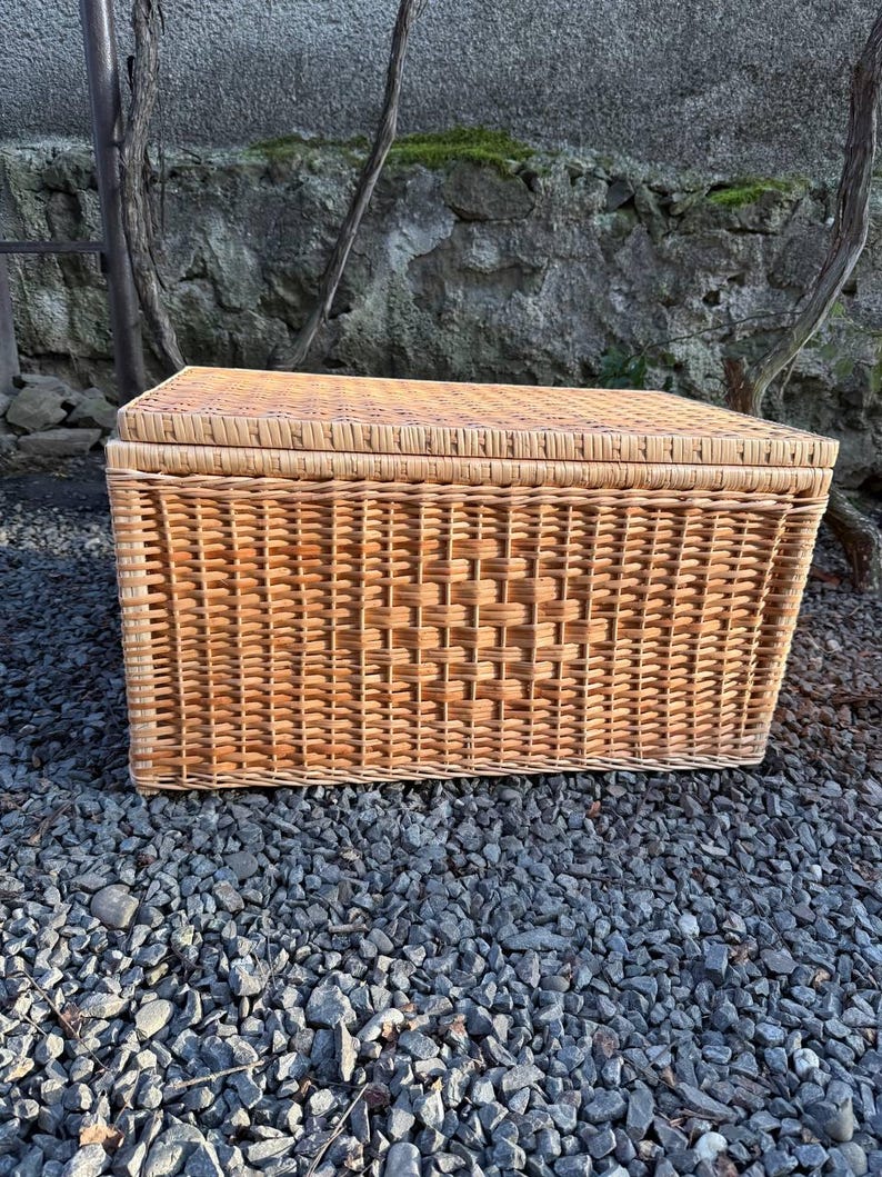 May include: A rectangular wicker storage basket with a lid. The basket is made of woven light brown wicker and sits on a bed of gray gravel. The basket has a woven pattern on the front.