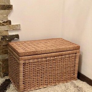 Puede incluir: Una cesta de almacenamiento de mimbre tejido con tapa. La cesta es de color marr&oacute;n claro y tiene un patr&oacute;n tejido natural. La cesta est&aacute; sentada sobre una alfombra marr&oacute;n claro.