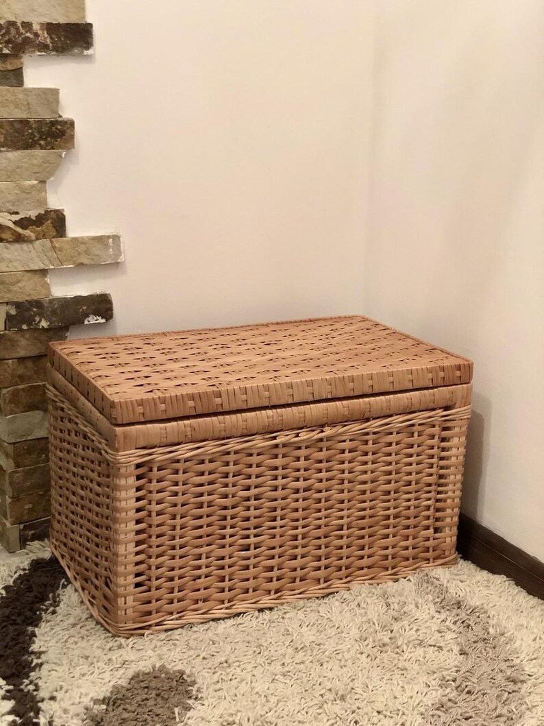 Puede incluir: Una cesta de almacenamiento de mimbre tejido con tapa. La cesta es de color marr&oacute;n claro y tiene un patr&oacute;n tejido natural. La cesta est&aacute; sentada sobre una alfombra marr&oacute;n claro.