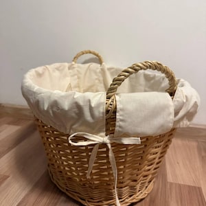 Puede incluir: Una cesta redonda de mimbre tejido, con un forro de tela color crema y asas de cuerda. La cesta es de color marrón claro y la tela crema está atada con un lazo. Ideal para el almacenamiento y la organización.