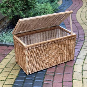 May include: A large, natural woven wicker storage basket with a hinged lid. The lid is open and the basket is sitting on a brick patio.