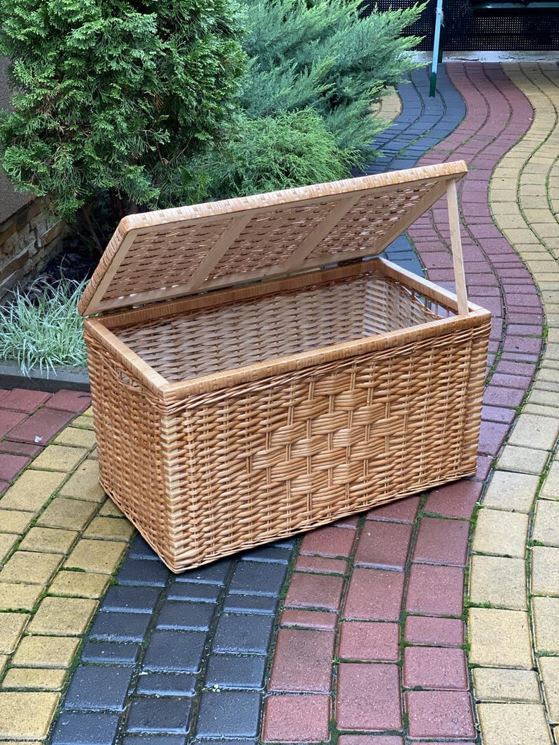 May include: A large, natural woven wicker storage basket with a hinged lid. The lid is open and the basket is sitting on a brick patio.