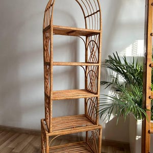 May include: A tall, arched, natural woven rattan shelf unit with four shelves. The shelf unit is made of natural woven rattan and has a light brown color. The shelf unit is standing on a wooden floor.