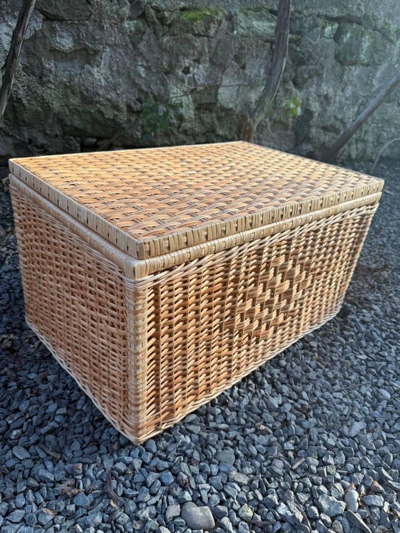 May include: A rectangular, light brown wicker storage basket with a matching lid. The basket is made of woven wicker and sits on a bed of small, dark gray stones. The basket is designed for storage and organization.