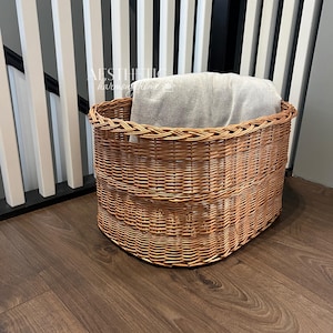 May include: A woven wicker basket with a gray blanket inside. The basket is oval shaped and has a natural brown color. The basket is sitting on a wooden floor.
