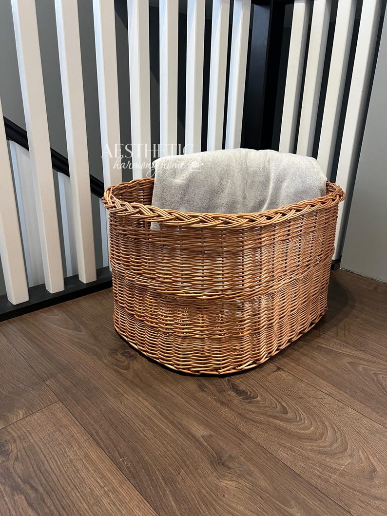 May include: A woven wicker basket with a gray blanket inside. The basket is oval shaped and has a natural brown color. The basket is sitting on a wooden floor.