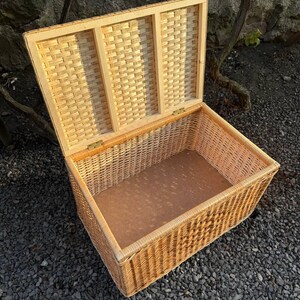 May include: An open rectangular wicker storage basket with a hinged lid. The basket is made of light brown woven wicker and has a light brown interior. The lid is partially open, revealing the interior. The basket is sitting on a bed of small, dark gray stones.