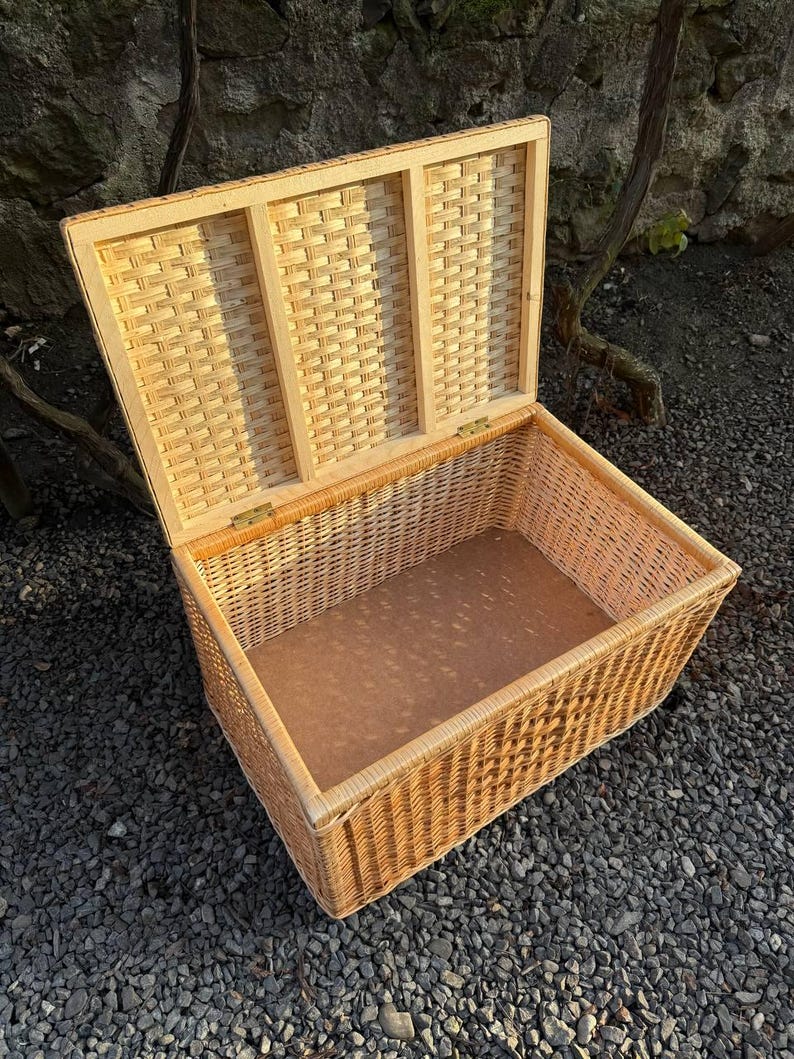May include: An open rectangular wicker storage basket with a hinged lid. The basket is made of light brown woven wicker and has a light brown interior. The lid is partially open, revealing the interior. The basket is sitting on a bed of small, dark gray stones.