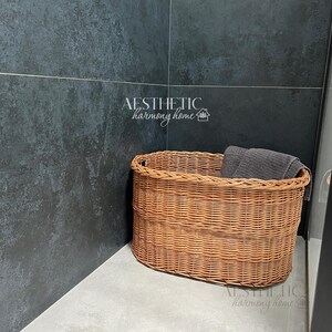May include: A brown wicker basket with a gray towel inside, sitting on a light gray tiled floor in a corner of a room with dark gray tile walls. The text "AESTHETIC harmony home" is visible on the wall.