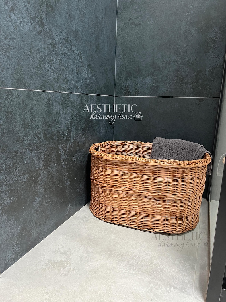 May include: A brown wicker basket with a gray towel inside, sitting on a light gray tiled floor in a corner of a room with dark gray tile walls. The text "AESTHETIC harmony home" is visible on the wall.