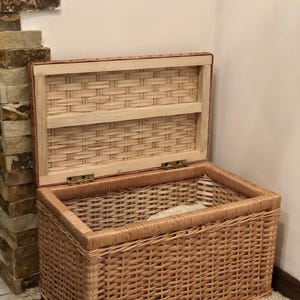 Puede incluir: Una cesta de almacenamiento de mimbre tejido con tapa con bisagras. La cesta es de color marr&oacute;n claro y tiene un acabado natural. La tapa est&aacute; abierta y revela un estante dentro de la cesta.