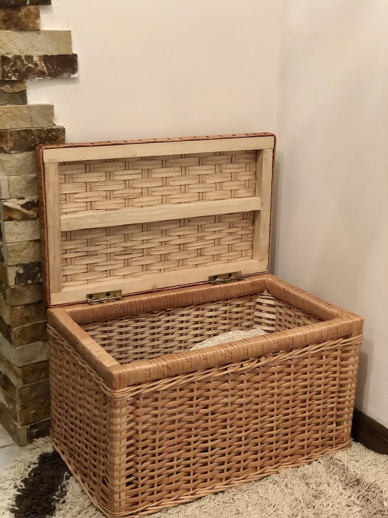 Puede incluir: Una cesta de almacenamiento de mimbre tejido con tapa con bisagras. La cesta es de color marr&oacute;n claro y tiene un acabado natural. La tapa est&aacute; abierta y revela un estante dentro de la cesta.