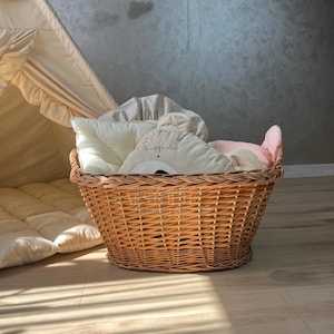 May include: A woven wicker basket filled with soft, light-colored pillows and bedding. A small, beige teepee is visible in the background, along with a cloud mobile. The basket is on a light wood floor, creating a cozy nursery scene.