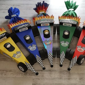 Könnte beinhalten: Fünf Pappkartons in Form von Rennwagen mit Flammen und karierten Flaggen. Jede Box hat eine andere Farbe: Gelb, Blau, Silber, Grün und Rot. Die Boxen haben ein schwarzes Namensschild mit der Aufschrift "Name" auf jeder.
