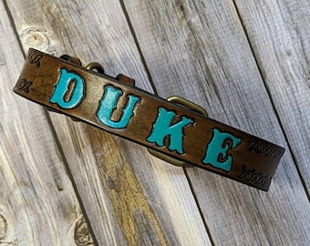 Puede incluir: Un collar de perro de cuero marrón con el nombre "DUKE" grabado en letras azul turquesa. El collar tiene una hebilla de latón y costuras decorativas.