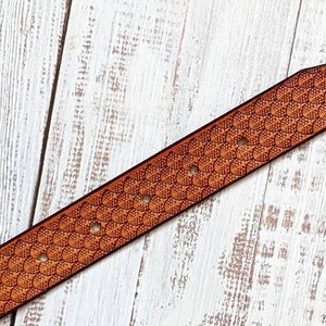 May include: A brown leather belt featuring a detailed scale pattern. The belt has several holes for adjustment and a tapered end. The design is embossed into the leather, creating a textured surface. The belt is set against a white wooden background.