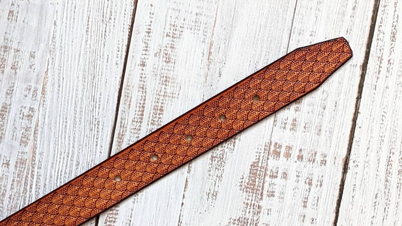 May include: A brown leather belt featuring a detailed scale pattern. The belt has several holes for adjustment and a tapered end. The design is embossed into the leather, creating a textured surface. The belt is set against a white wooden background.
