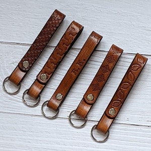 May include: Six brown leather keychains with different designs and metal key rings. The designs include a geometric pattern, a floral pattern, a word, and a leaf pattern.