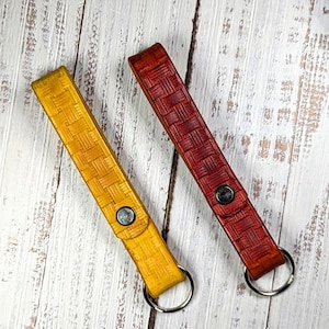 May include: Two leather keychains with a woven pattern. One is a golden yellow, the other is a deep red. Each keychain has a metal snap and a silver key ring. The keychains are displayed on a light wood surface.
