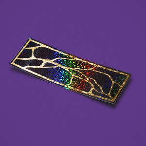 Op de afbeelding: Een zwarte en gouden iriserende sticker met een regenboogpatroon en een boomvormig ontwerp.
