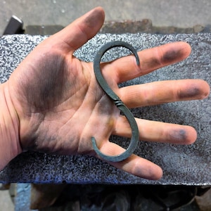 Peut inclure: Une main tenant un crochet en métal gris foncé en forme de S. Le crochet est en métal torsadé et a une finition lisse.