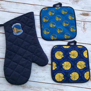 May include: Set of three Golden State Warriors themed kitchen accessories. A blue oven mitt with a Golden State Warriors logo and two blue and yellow pot holders with a basketball and team logo pattern.