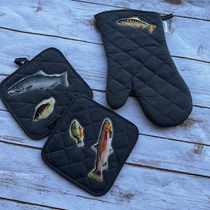 May include: A set of dark gray quilted kitchen accessories featuring fish designs. The set includes an oven mitt and two square pot holders. The fish illustrations are in various colors, including silver, orange, and yellow.