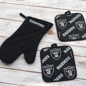 May include: Two black oven mitts with a quilted design. One mitt is a standard oven mitt and the other is a smaller pot holder. Both mitts feature a repeated pattern of the Las Vegas Raiders logo and the word "RAIDERS" in silver.