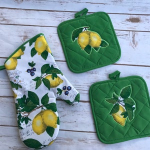May include: A set of two green lemon-themed pot holders and a white lemon-themed oven mitt. The pot holders have a quilted texture and the oven mitt has a white background with a lemon and leaf pattern.