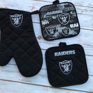 May include: Set of three black quilted oven mitts and pot holders with the Las Vegas Raiders logo. The logo features a silver helmet with crossed swords and the word "RAIDERS" in silver lettering.