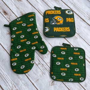 May include: Green and gold oven mitts and pot holders with the Green Bay Packers logo and the word "Packers" repeated on the fabric.