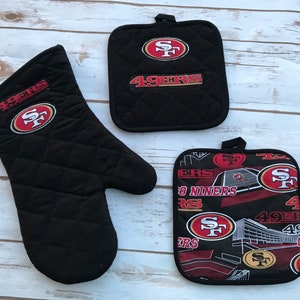 May include: A black quilted oven mitt and two pot holders featuring the San Francisco 49ers logo. The oven mitt and one pot holder have the team logo and "49ERS" in red and gold. The other pot holder has a repeating pattern.