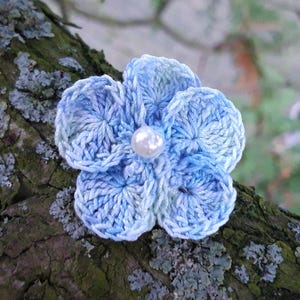 Peut inclure: Une fleur au crochet bleu clair faite à la main avec un centre de perle. La fleur est faite de fil doux et a cinq pétales. La fleur est fixée à une branche d'arbre.