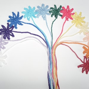 May include: A selection of handmade crochet flowers in a rainbow of colours, including blue, purple, green, pink, yellow, and orange. Each flower has a long stem, creating a vibrant and artistic display against a white background.