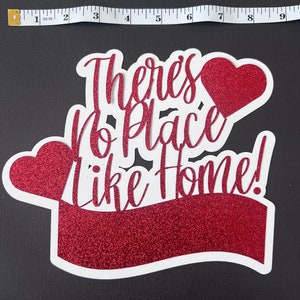 May include: Red glitter cake topper with the text "There's No Place Like Home!" and two red glitter hearts.