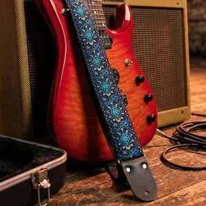 Red electric guitar leaning on a tweed amp displaying a blue floral guitar strap. High-quality handmade gear with leather ends for studio setups.