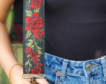 May include: A black crossbody bag with a floral strap. The strap has red and green embroidered flowers. The bag is being held by a person wearing blue jeans.