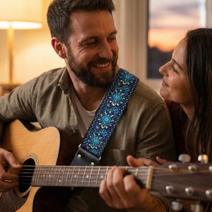 Man playing acoustic guitar at home using a blue patterned strap. Comfortable handmade accessory perfect for cozy indoor practice sessions.