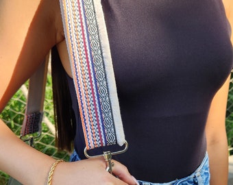 Pode incluir: Um top preto sem mangas com uma alça estampada em branco e colorido. A alça está presa a uma bolsa de couro preta com tachas prateadas. A pessoa está usando jeans azul e um colar de pérolas.