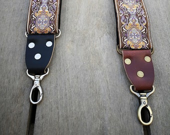 Op de afbeelding: Twee gitaarbanden met een bruin en gouden patroon. De band aan de linkerkant heeft een zwart leren uiteinde met witte stippen en een zilveren haak. De band aan de rechterkant heeft een bruin leren uiteinde met messing stippen en een messing haak.