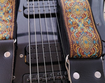 May include: A black electric guitar with a decorative guitar strap. The strap features a brown, teal, and gold ornate pattern. The strap is attached to the guitar with black leather ends and silver rivets.