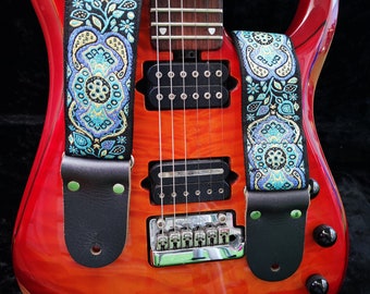 May include: A red electric guitar with a black and blue patterned guitar strap. The strap has a leather end with two green dots.
