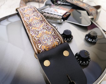 May include: A brown guitar strap with a colorful floral pattern and a black leather end. The strap is attached to a silver electric guitar.