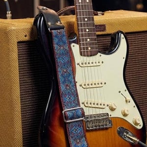 May include: A sunburst electric guitar with a white pickguard and a blue patterned strap leans against a vintage-style amplifier. A coiled cable connects the guitar to a green effects pedal.