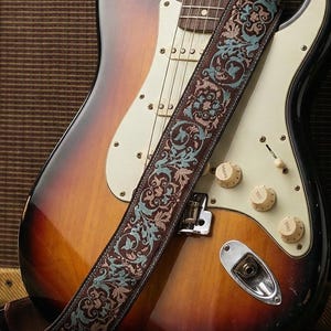 Może przedstawiać: Sunburst gitara elektryczna z ozdobnym paskiem opiera się o wzmacniacz w stylu vintage. Gitara ma biały pickguard i kremowe pokrętła. Zwinięty kabel leży na drewnianej podłodze. Druga gitara jest widoczna w tle.